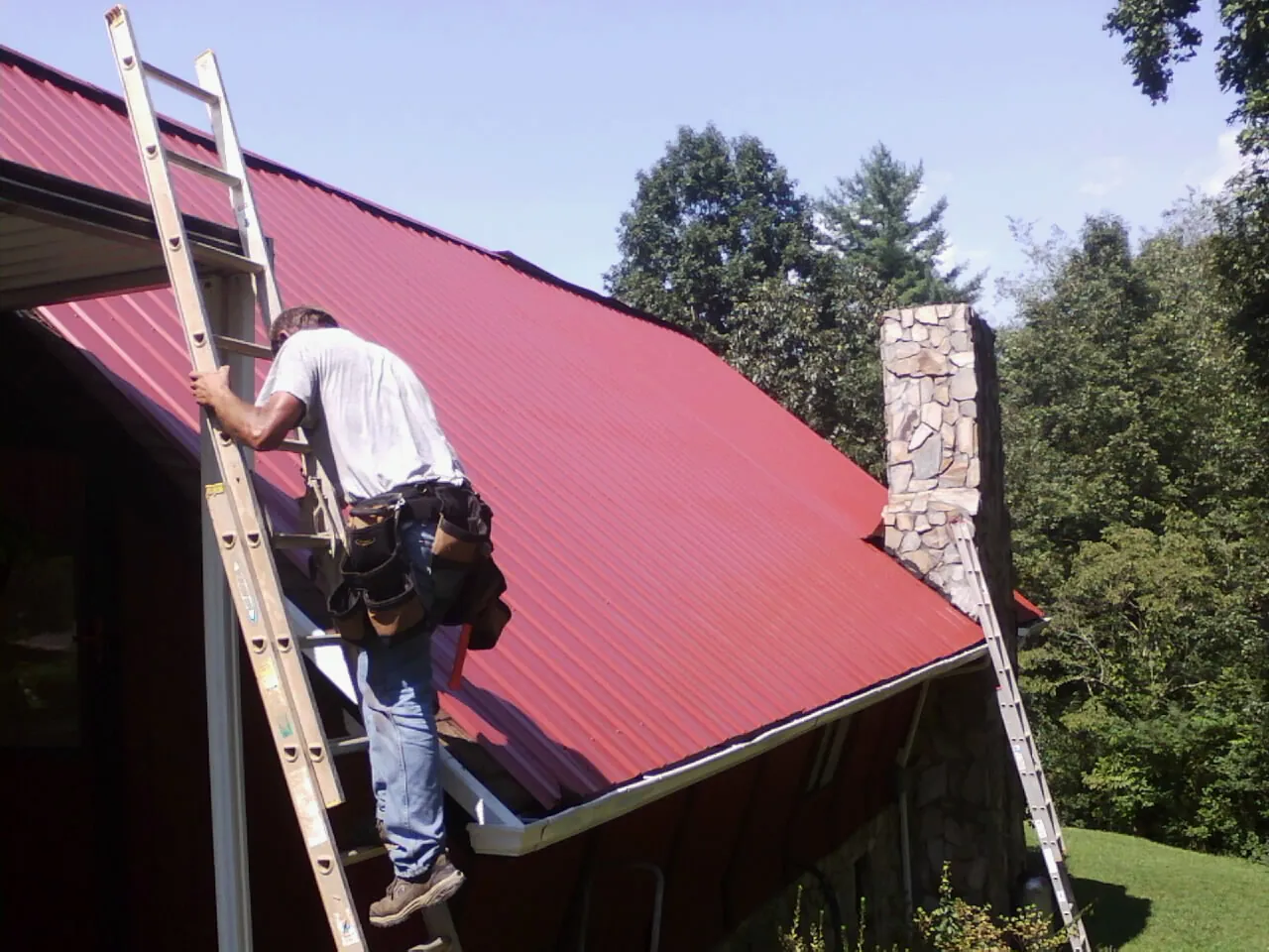 Roofer on a ladder beside a red metal roof and stone chimney — Soffit & Fascia Repair by Roofing Brothers in Thayne